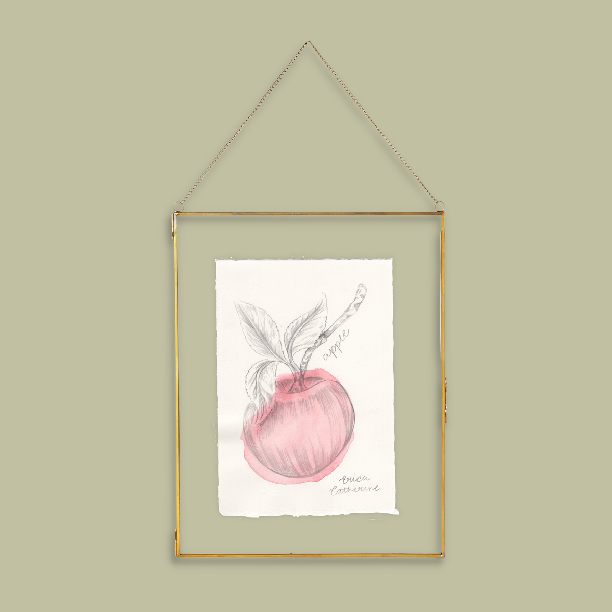 A framed botanical art print of an apple hanging on a light olive green wall. The print features a pink watercolor apple with a pencil sketch of leaves and a stem, set in a thin gold frame with a hanging chain.