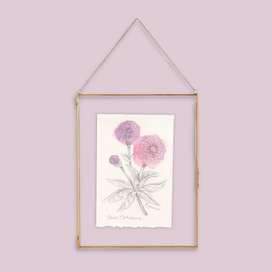 A gold-framed botanical art print featuring pink and purple Zinnia flowers on a white background, suspended by a thin gold chain against a solid light pink wall.