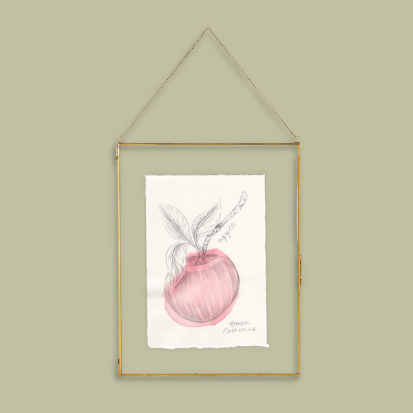 A framed botanical art print of an apple hanging on a light olive green wall. The print features a pink watercolor apple with a pencil sketch of leaves and a stem, set in a thin gold frame with a hanging chain.