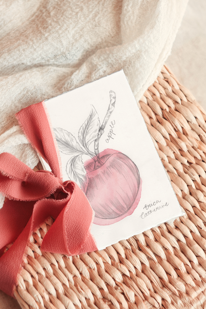 An artwork featuring a watercolor sketch of an apple, tied with a coral pink ribbon and displayed on a woven wicker basket with a soft cream textile background.