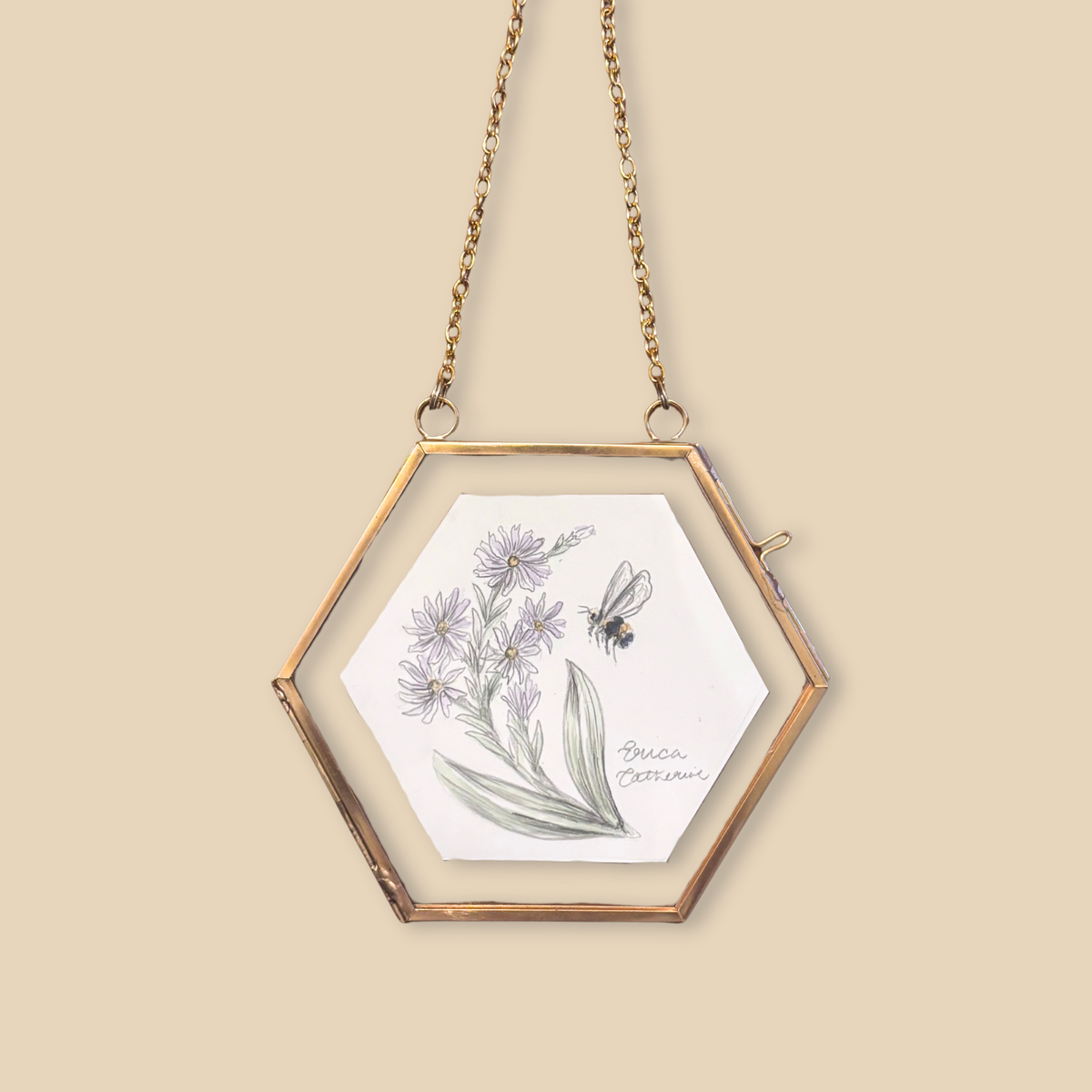 A hexagonal hanging frame with a gold-toned metal border and chain, containing a white card with a botanical illustration of purple flowers and a bee, set against a solid beige background.