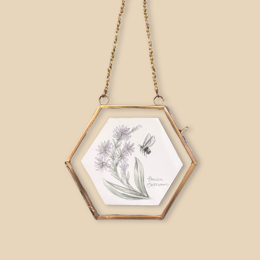 A hexagonal hanging frame with a gold-toned metal border and chain, containing a white card with a botanical illustration of purple flowers and a bee, set against a solid beige background.