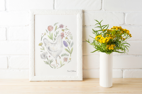 Framed artwork of a chicken and flowers next to a vase with yellow flowers on a wooden surface.