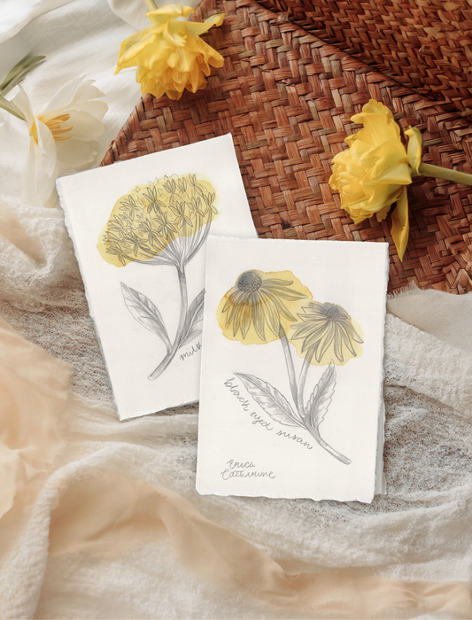 Two botanical art prints featuring yellow flowers, styled on a neutral fabric surface next to a woven basket and yellow flowers.