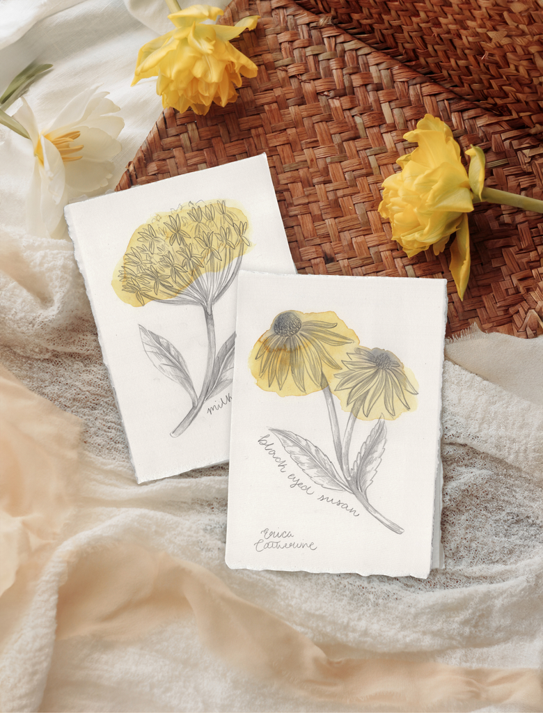 Two botanical art prints featuring yellow flowers, styled on a neutral fabric surface next to a woven basket and yellow flowers.