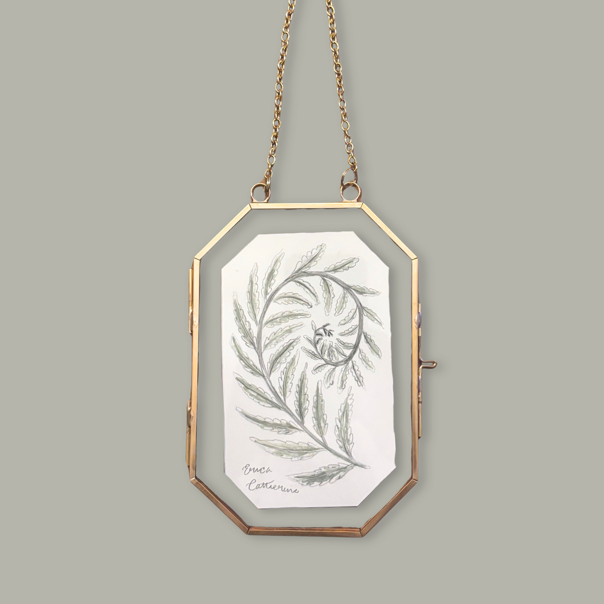 A gold-framed octagonal hanging clutch bag with a white interior card featuring a botanical illustration, suspended against a solid gray background.