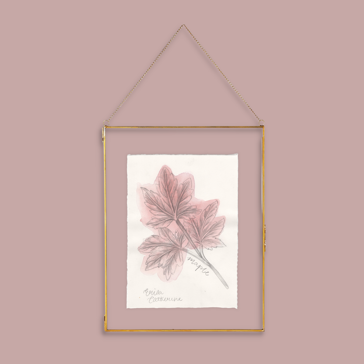 A gold-framed hanging wall art print featuring a botanical illustration of pinkish maple leaves on textured white paper, suspended by a gold chain against a dusty pink background.