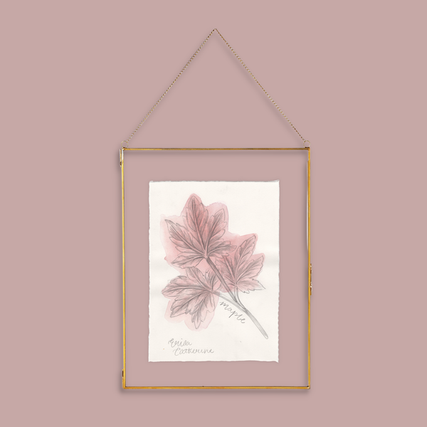 A gold-framed hanging wall art print featuring a botanical illustration of pinkish maple leaves on textured white paper, suspended by a gold chain against a dusty pink background.