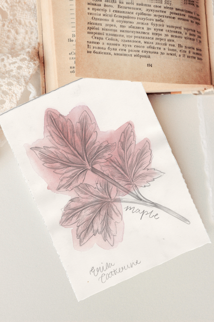 An artwork featuring a watercolor illustration of a maple leaf in dusty pink and gray, with the word 'maple' and Erica Catherine's signature styled on top of an open vintage book.