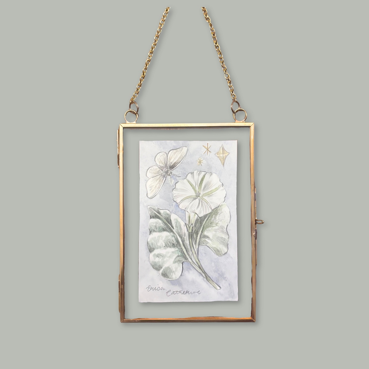 A rectangular hanging art frame with a thin gold-toned border and chain, containing a botanical illustration of white flowers and green leaves on a pale background, shown against a solid gray backdrop.