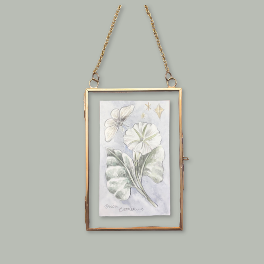 A rectangular hanging art frame with a thin gold-toned border and chain, containing a botanical illustration of white flowers and green leaves on a pale background, shown against a solid gray backdrop.