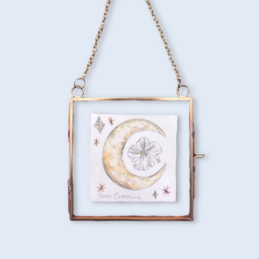 A square hanging frame with a gold-toned metal border and chain, featuring a central white card with a watercolor-style crescent moon, flower, and small stars. The frame is shown against a pale blue background.