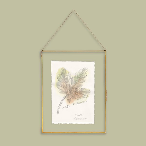 A botanical art print of an oak leaf and acorn, presented in a thin gold metal frame with a hanging chain against a solid sage green background.