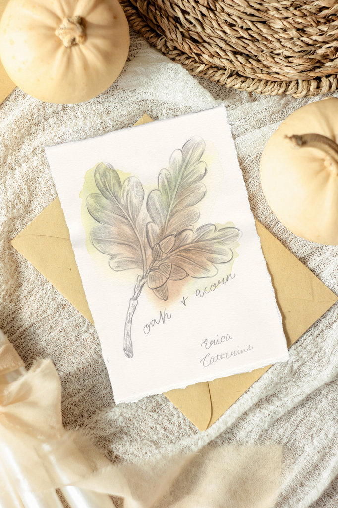 A flat-lay of an artwork featuring a watercolor oak leaf and acorn illustration with the text 'oak + acorn' and Erica Catherine's Signature. The print is styled on a textured neutral fabric with kraft paper envelopes, autumn pumpkins, and a woven basket.