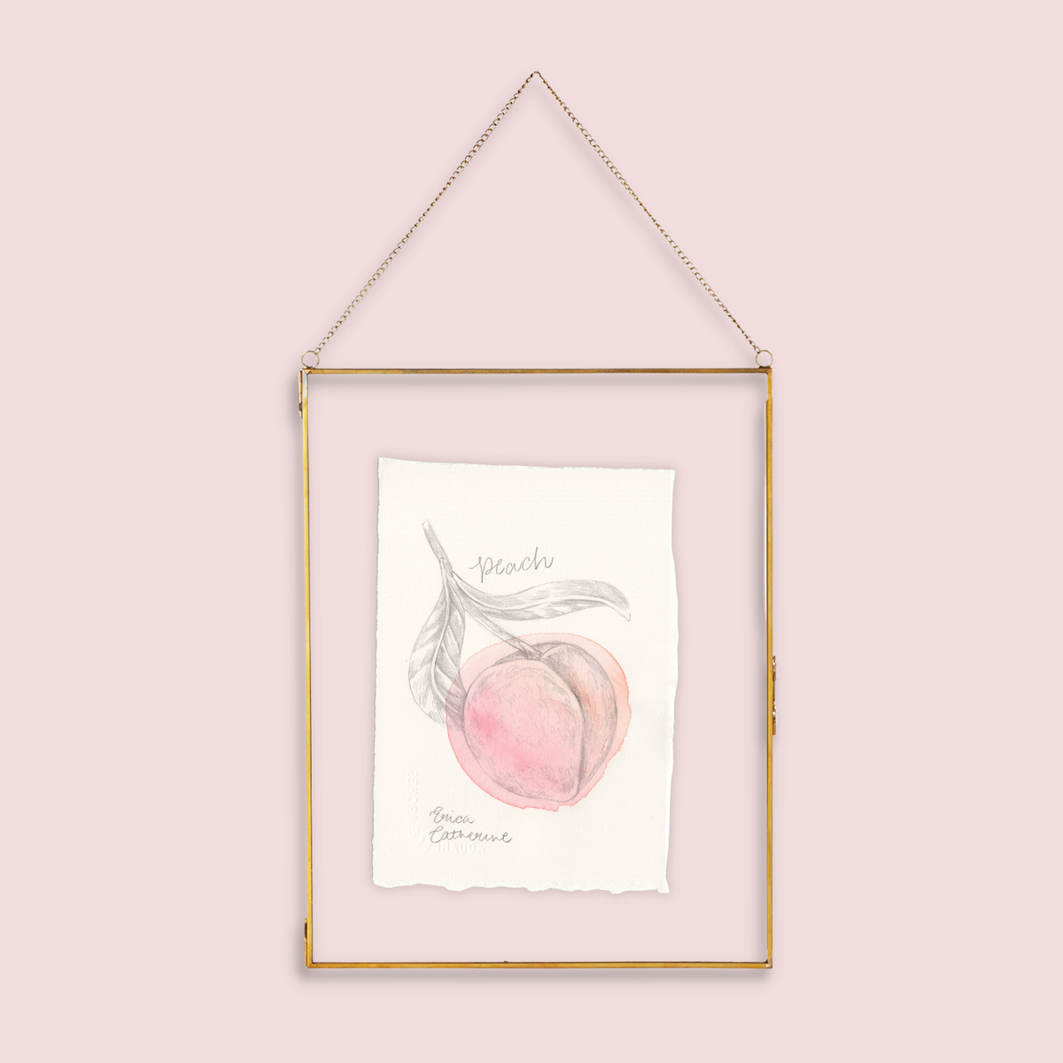 A gold-framed hanging art print featuring a watercolor illustration of a peach with the word 'Peach' written below, suspended against a solid pale pink background.
