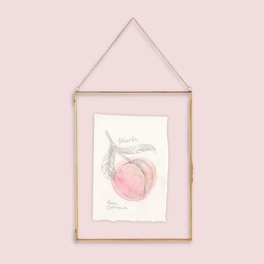 A gold-framed hanging art print featuring a watercolor illustration of a peach with the word 'Peach' written below, suspended against a solid pale pink background.