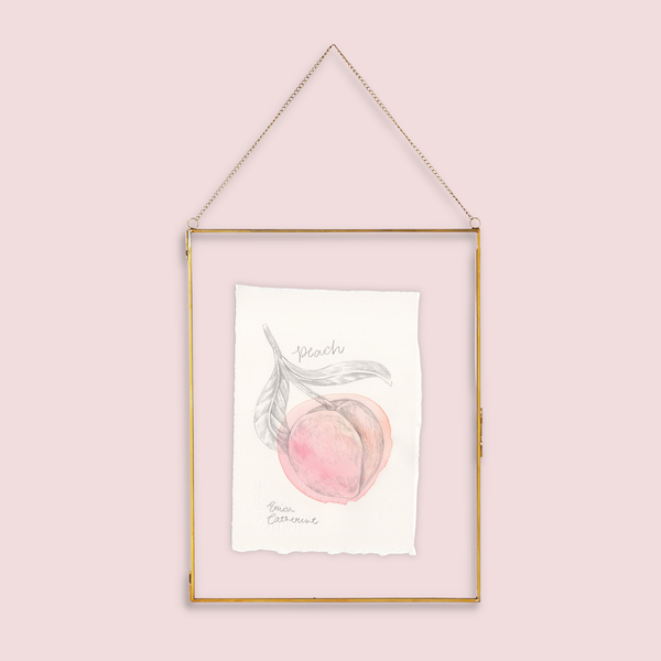 A gold-framed hanging art print featuring a watercolor illustration of a peach with the word 'Peach' written below, suspended against a solid pale pink background.