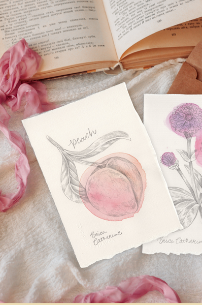 Two small artworks with watercolor illustrations of peaches and flowers, styled on a cream fabric with an open book and pink ribbon in the background.