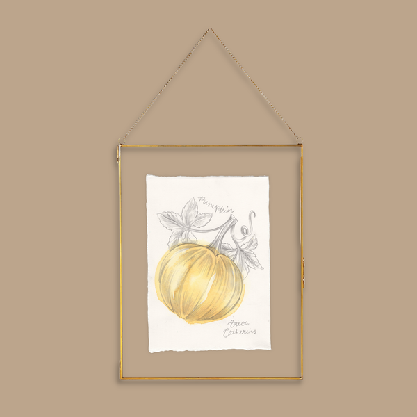 A framed artwork featuring a watercolor pumpkin illustration on a white sheet of paper, enclosed in a thin gold metal frame with a hanging chain, set against a solid beige background.
