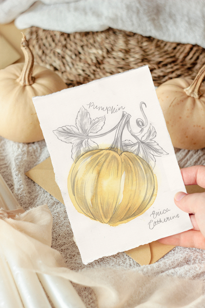 A hand holding an artwork featuring a painted yellow pumpkin with gray botanical details, styled with real pumpkins, fabric, and paper in a warm autumn setting.
