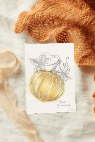 A small artwork featuring a hand-drawn yellow pumpkin with gray leaves, styled on a white fabric background with an orange knit blanket and beige ribbon.