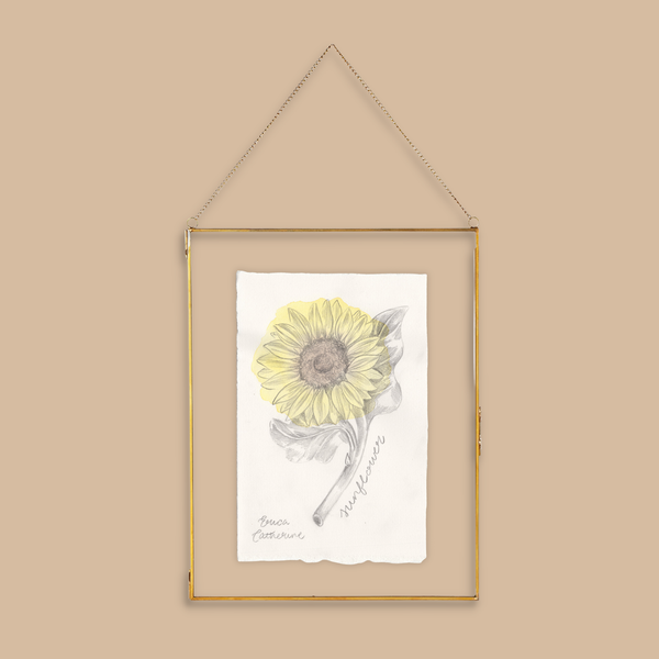 A gold-framed hanging art print featuring a watercolor-style yellow sunflower on white paper, suspended by a thin gold chain against a solid beige background.