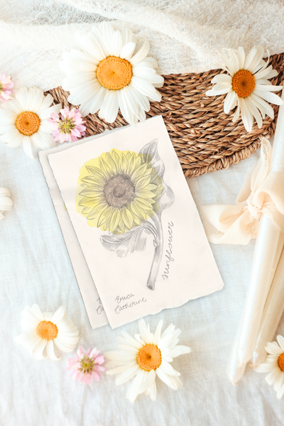A flat-lay photograph of a vintage-style botanical artwork featuring a watercolor sunflower, surrounded by white and pink daisies, a woven straw basket, and cream-colored candles on a white textured fabric background.